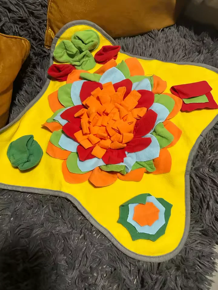Buyer photo: the Sunny yellow snuffle mat on a gray rug, orange flower core with red, green and pale blue petals and a gray trim